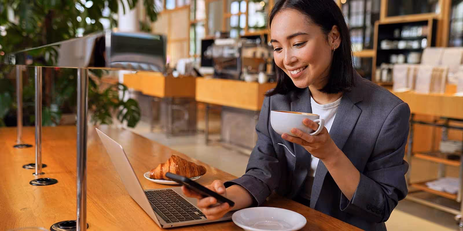 Woman smiling while working remotely in a café, holding a cup of coffee and looking at her phone with a laptop and croissant on the table