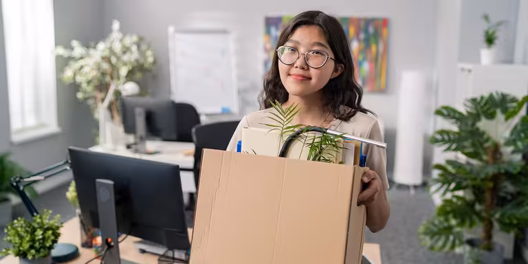 Smiling woman in an office, holding a box of her belongings, leaving her job after quitting