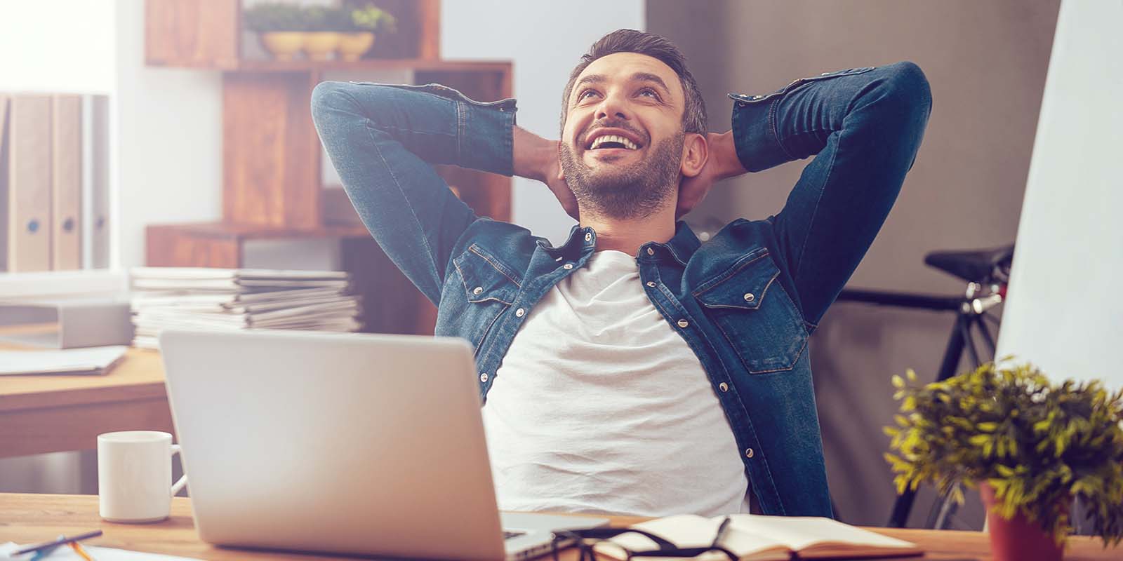 Man sitting at a desk with a laptop, leaning back with hands behind his head and smiling, appearing relaxed and content in an office setting