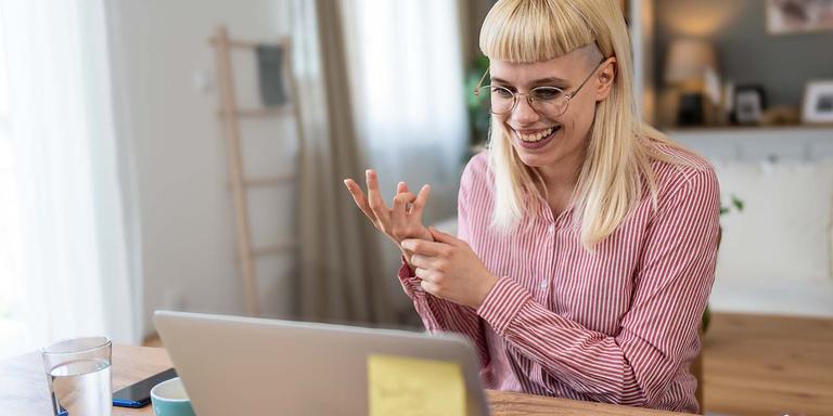 Smiling person with long blonde hair and glasses sitting at a desk, gesturing while on a video call using a laptop in a bright home setting