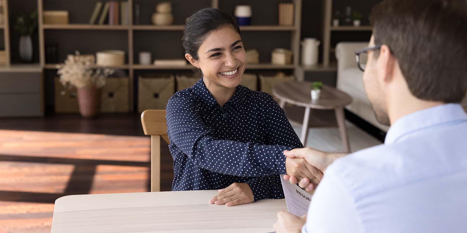 Smiling job candidate shakes hands with interviewer holding a resume across a table, indicating she was hired after a successful interview