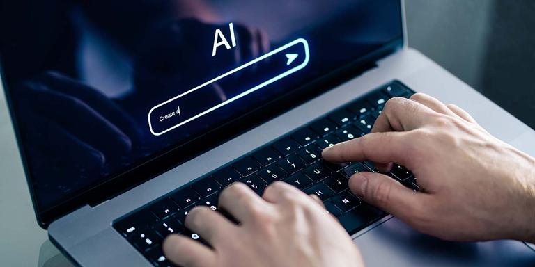 A close-up view of a person’s hands typing on a laptop keyboard. The laptop screen displays an AI interface with a search bar that contains the text "Create a|" and an "AI" logo above it.