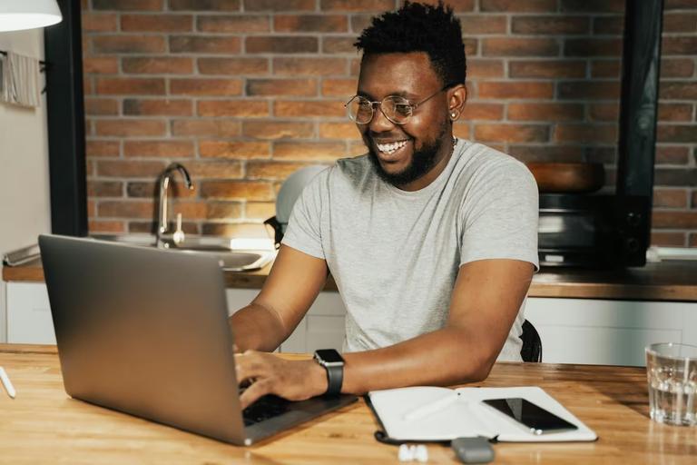 Smiling man works on a laptop at a wooden table in a modern kitchen, with a notebook and smartphone nearby, illustrating a job seeker searching for a new job online in 2026.